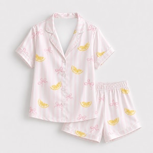 Op de afbeelding: Een tweedelige pyjamaset bestaande uit een shirt met korte mouwen en knoopsluiting en een bijpassende short. De stof is lichtroze met witte verticale strepen, versierd met een herhalend patroon van gele citroenschijfjes en kleine roze strikjes.