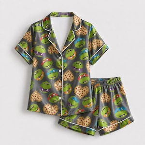 May include: A dark gray pajama set consisting of a short-sleeved button-down shirt and matching shorts. The fabric features a repeating pattern of green cartoon turtle faces with various colored masks and tan heart-shaped pizza slices.