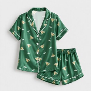 Op de afbeelding: Een donkergroene pyjamaset bestaande uit een overhemd met korte mouwen, knoopsluiting en reverskraag, en een bijpassende short. De stof heeft een all-over print van martiniglazen met olijven, afgewerkt met witte biezen langs de randen.