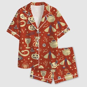 Puede incluir: Un conjunto de pijama de camisa de manga corta con botones y pantalón corto a juego en color rojo oscuro. La tela presenta un patrón repetitivo de ilustraciones de color crema con temas ocultistas y de Halloween, incluyendo gatos, fantasmas, calabazas, polillas y calderos.