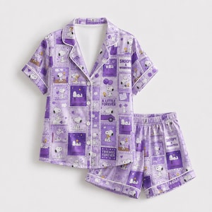 May include: A purple pajama set consisting of a short-sleeved button-down shirt and matching shorts. The fabric features a repeating grid pattern of Snoopy and Woodstock illustrations, with text including "Snoopy & Woodstock" and "Friends Forever."