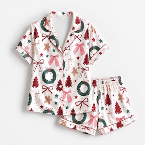 May include: A white short-sleeve pajama set featuring a festive holiday print of green wreaths, red Christmas trees, pink and red bows, gingerbread cookies, and small green stars. The button-down shirt has a notched collar and red piping.