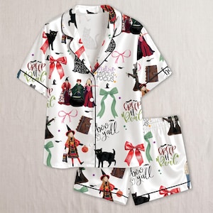 May include: A white pajama set with short sleeves and shorts featuring a repeating pattern of black cats, red bows, green ribbons, spellbooks, and three witch figures. The text reads Creep it real, It's just Hocus Pocus, and Boo y'all.