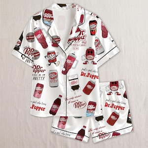 May include: A white short-sleeve button-down pajama shirt and matching shorts set featuring an all-over print of Dr. Pepper soda cans, tumblers, and bottles. The pattern includes the text Dr. Pepper and the phrase Just a girl who loves Dr. Pepper.