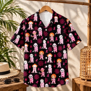 Puede incluir: Una camisa negra de manga corta con botones y un estampado integral de varios personajes de películas de terror vestidos con trajes rosas, intercalados con telarañas rosas y pequeños iconos de lazos rosas. La camisa tiene cuello y botones blancos.