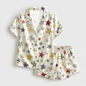 May include: A two-piece pajama set featuring a short-sleeved button-down shirt with a notched collar and matching elastic-waist shorts. Both pieces are made of a cream-colored fabric covered in a repeating pattern of multi-colored stars.