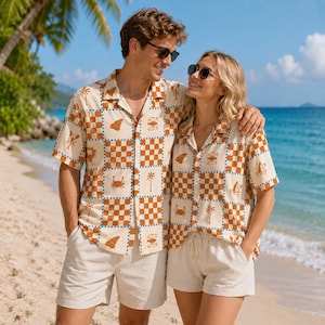 May include: Two short-sleeved button-down shirts featuring a cream and tan checkered pattern with small brown illustrations of palm trees, crabs, seashells, fish, and sailboats. The shirts have a classic collar and are paired with beige shorts.