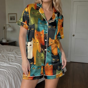 May include: A short-sleeve button-down pajama shirt and matching shorts set featuring a vibrant, all-over print of stylized cats in various colors including orange, blue, green, black, and white, finished with white piping along the edges.