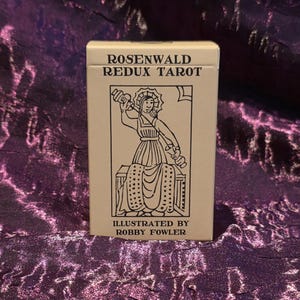 May include: A beige tarot card box featuring the text "ROSENWALD REDUX TAROT" and an illustration of a woman. The box also includes the text "ILLUSTRATED BY ROBBY FOWLER". The background is a dark purple fabric.