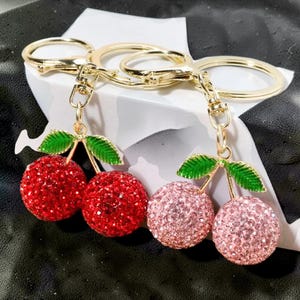 May include: Two keychains with gold-colored rings and clasps. Each keychain features a pair of cherry charms. One pair is red, and the other is pink. The cherries are covered in sparkling rhinestones, with green leaves.