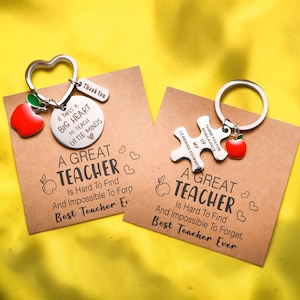 Op de afbeelding: Twee sleutelhangers voor leraren, elk met een rode appelbedel. De ene sleutelhanger heeft een hartvormige ring en een schijfje met de tekst "It takes a big heart to teach little minds". De andere heeft een puzzelstukje met "Thank you for being a piece of my learning journey."