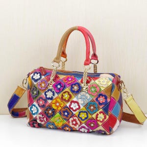 May include: A multi-coloured handbag featuring a diamond-patterned exterior with attached leather flowers. Each flower has a small clear rhinestone at its centre. The bag includes two top handles in tan and red, and a detachable shoulder strap.