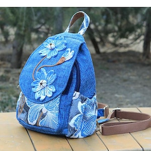 May include: A blue denim backpack featuring a top flap adorned with two three-dimensional fabric flowers. The lower section and side pockets display a white and blue floral pattern. The bag includes a top handle and adjustable brown fabric straps.
