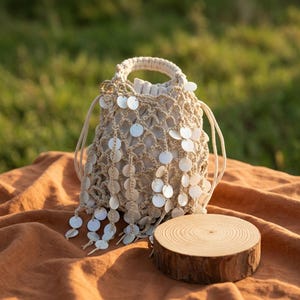 May include: A beige macrame bucket bag featuring a drawstring closure and a net-like exterior overlay. The bag is adorned with numerous small, round, white shell-like discs that hang from the netting, creating a textured and layered appearance.