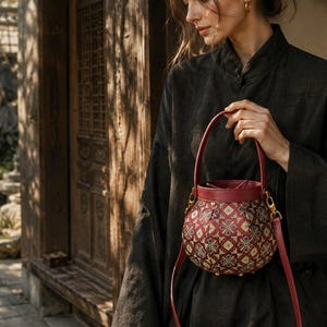 May include: A round bucket bag featuring a deep red leather top handle and trim, paired with a patterned fabric body displaying a geometric floral motif in shades of red, gold, and teal. The bag includes a detachable red leather shoulder strap.
