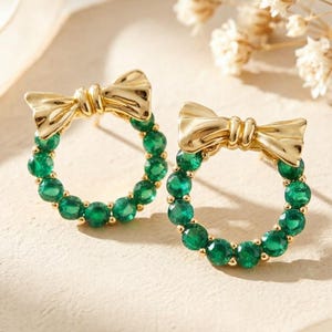 May include: A pair of gold-tone stud earrings featuring a circular hoop design adorned with small, round, vibrant green stones. Each earring is topped with a polished gold-tone bow accent, creating a cohesive look with the circular gemstone frame.