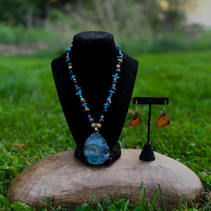 Può includere: Una collana con un grande ciondolo in pietra blu a forma di goccia sospeso a un filo di perline turchesi, marroni e nere, accompagnata da un paio di orecchini in pietra marrone a forma di goccia su un espositore nero.