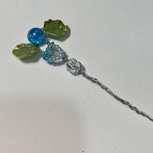 May include: A handmade hair accessory with a floral design. It features a blue bead, green leaves, and a heart-shaped cluster of small beads. The stem is made of twisted silver wire.