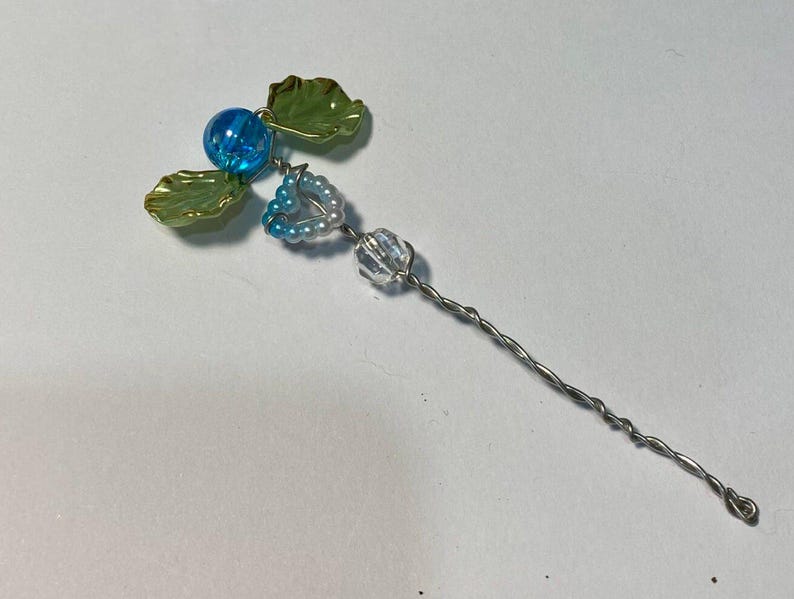 May include: A handmade hair accessory with a floral design. It features a blue bead, green leaves, and a heart-shaped cluster of small beads. The stem is made of twisted silver wire.