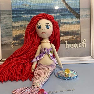 May include: A handmade mermaid doll with long red yarn hair, a pink and white patterned tail, and a purple top. A small tiara and a decorative wand are next to the doll. The word "beach" is visible in the background.