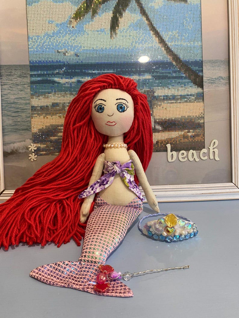 May include: A handmade mermaid doll with long red yarn hair, a pink and white patterned tail, and a purple top. A small tiara and a decorative wand are next to the doll. The word "beach" is visible in the background.