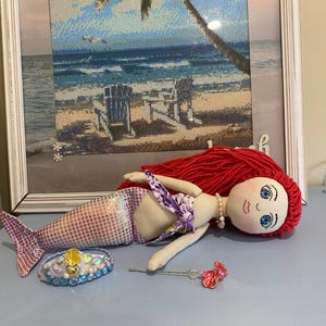 May include: A handmade mermaid doll with red yarn hair, blue eyes, and a pearl necklace. The doll has a purple and white floral top and a patterned tail. A bejeweled crown and a pink and clear wand are next to the doll.