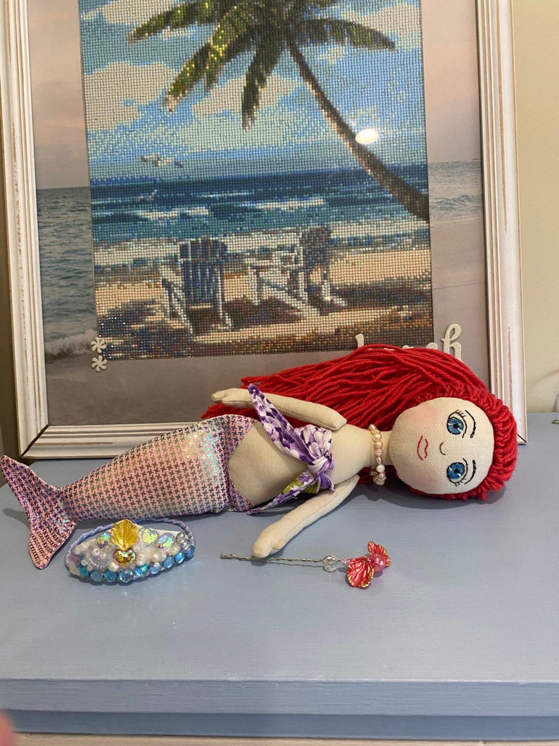 May include: A handmade mermaid doll with red yarn hair, blue eyes, and a pearl necklace. The doll has a purple and white floral top and a patterned tail. A bejeweled crown and a pink and clear wand are next to the doll.