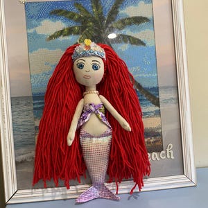 May include: A handmade mermaid doll with red yarn hair, a fabric face, and a shimmering purple tail. The doll wears a floral top and a crown. A small wand with a pink bow lies nearby. The doll is displayed in a white framed picture with a beach scene.