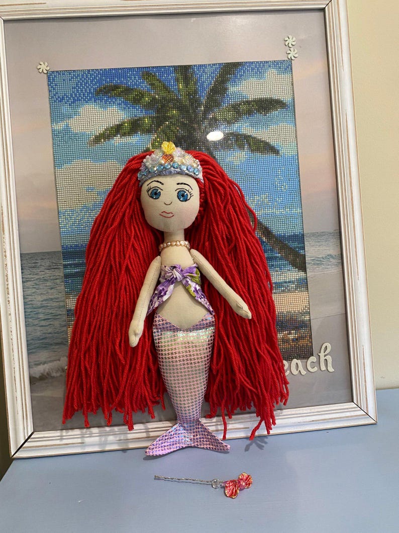 May include: A handmade mermaid doll with red yarn hair, a fabric face, and a shimmering purple tail. The doll wears a floral top and a crown. A small wand with a pink bow lies nearby. The doll is displayed in a white framed picture with a beach scene.