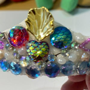 May include: A decorative tiara with a white base, adorned with iridescent gems, pearls, and a gold leaf accent. The tiara features a variety of colourful, faceted stones in shades of blue, red, and green, creating a sparkling effect.