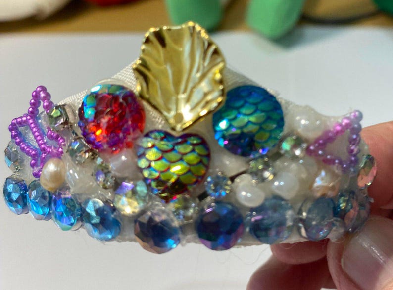May include: A decorative tiara with a white base, adorned with iridescent gems, pearls, and a gold leaf accent. The tiara features a variety of colourful, faceted stones in shades of blue, red, and green, creating a sparkling effect.