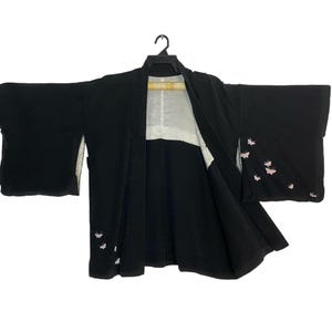 May include: A black haori jacket featuring wide sleeves and a white inner lining. The exterior is decorated with small, light pink butterfly motifs embroidered on the lower front panel and the outer surface of the right sleeve.