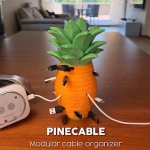 May include: A pineapple-shaped orange and green modular cable organizer labeled PINECABLE. The device features multiple slots around its body, each holding a black or white charging cable connector, with one cable plugged into a nearby electronic device.