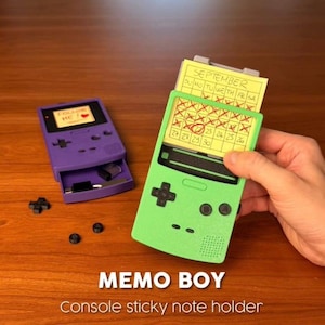 May include: A purple and a light green handheld game console shaped sticky note holder. The green holder displays a yellow September calendar sticky note with handwritten dates. The purple holder has a small drawer containing small items.