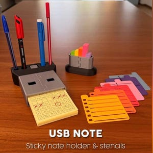 May include: A black USB-shaped desk organiser with pens, sticky notes, and stencils. The organiser holds a calendar notepad and colourful sticky notes. The text "USB NOTE" and "sticky note holder & stencils" are visible.