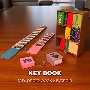 May include: A brown miniature bookshelf containing colorful square keychains, two extended photo book strips, and two closed square keychains with the text KEY BOOK Mini photo book keychain printed below the items.