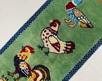May include: A rectangular green runner rug featuring a repeating pattern of colourful roosters and birds. The rug has a dark blue and black textured border. The birds are depicted in shades of red, yellow, blue, white, and brown against the green field.