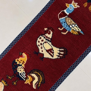 May include: A long, rectangular runner rug featuring a deep red background with a repeating pattern of colorful birds, including roosters and ducks, rendered in a pixelated style. The rug is framed by a narrow, dark blue and black checkered border.