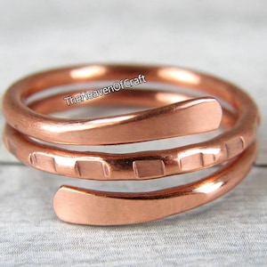 May include: A copper ring featuring a spiral design with three overlapping bands. The middle band displays a series of small, evenly spaced indentations along its surface. The text TheHeavenOfCraft is visible on the upper band of the ring.