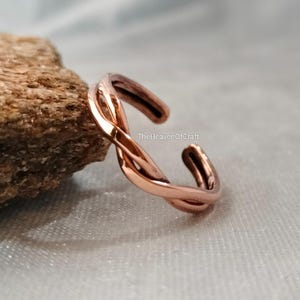 May include: A copper-coloured metal ring featuring a twisted, open-band design. The surface of the ring has a polished, reflective finish. The text TheHeavenOfCraft is visible on the inner curve of the band, which is open at one end.