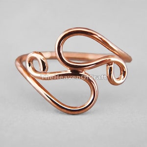 Può includere: Un anello color oro rosa lucido caratterizzato da un design a filo continuo e fluido con molteplici anelli e curve aperte. Il testo TheHeavenOfCraft è sovrapposto al centro della fascia metallica, che presenta una superficie liscia e riflettente.