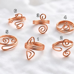 Peut inclure: Six bagues en fil de cuivre avec des anneaux ouverts et réglables, chacune présentant des motifs géométriques ou en spirale distincts étiquetés avec les chiffres 1 à 6. Les bagues sont formées de fil de cuivre poli et fin.
