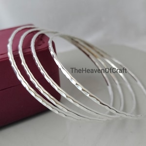 Puede incluir: Un conjunto de cuatro brazaletes finos de metal color plata con una superficie martillada y texturizada. Las pulseras están dispuestas en una pila, superponiéndose parcialmente, con el texto TheHeavenOfCraft visible en el centro.