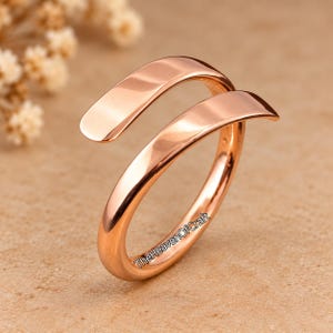 May include: A polished rose gold-tone bypass ring with a smooth, open-ended design. The metal surface is reflective and curved, featuring the text TheHeavenOfCraft printed in small black letters along the side of the band.