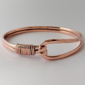 Puede incluir: Un brazalete de metal en tono oro rosa con un diseño de alambre liso y redondeado. La pulsera se asegura con un cierre de tipo gancho, donde un extremo del alambre está envuelto firmemente alrededor del otro en una serie de espirales paralelas.