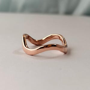 May include: A rose gold ring featuring a continuous, smooth, undulating wave design. The metal has a polished, reflective surface that catches the light, creating a fluid, curved shape that forms a single, unbroken band with a consistent width.