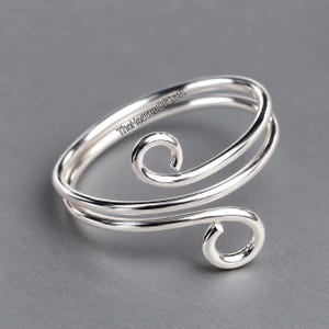 Op de afbeelding: Een zilverkleurige wikkelring met een dubbel draadontwerp en twee spiraalvormige uiteinden. Het metalen oppervlak is glad en reflecterend. De tekst TheHeavenOfCraft is in het zwart gedrukt op de bovenste draad van deze open ring.