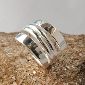 Könnte beinhalten: Ein polierter Silberring mit einem mehrschichtigen Wickeldesign, bei dem sich drei einzelne Bänder überlappen und einen durchgehenden Spiraleffekt erzeugen. Die Metalloberfläche ist glatt und reflektierend und fängt das Licht an den Kanten ein.