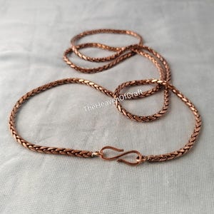 May include: A copper-coloured braided chain necklace featuring a metallic S-hook clasp. The chain has a woven, textured appearance and is laid out in a circular shape. The text TheHeavenOfCraft is visible in the centre of the chain loop.