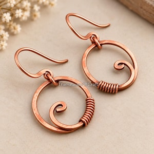 May include: A pair of copper earrings featuring a circular hoop design with a hammered texture. Each earring includes a central spiral element and a section of wire wrapping along the side, suspended from a curved ear wire hook. The text TheHeavenOfCraft is visible.